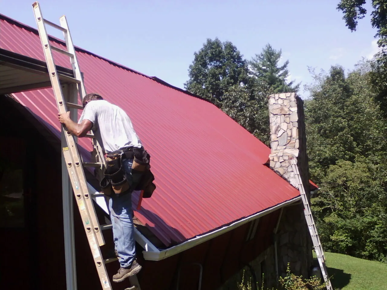Roofer on a ladder beside a red metal roof and stone chimney — Soffit & Fascia Repair by Roofing Brothers in Lacygne